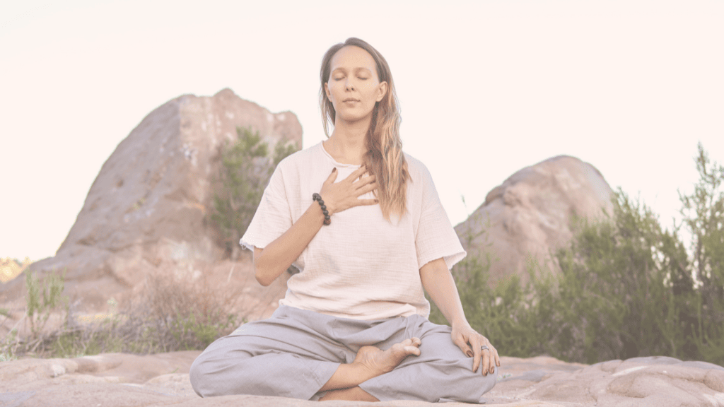 How Your Attachment Style in Relationships Lead to Divorce & 4 Ways to Create a More Secure Attachment Style | MCD woman with long blonde hair, white shirt and blue yoga pants, sitting in a yoga pose in the desert, hills behind her, eyes closed and the right hand on her heart, how to create a more secure attachment style