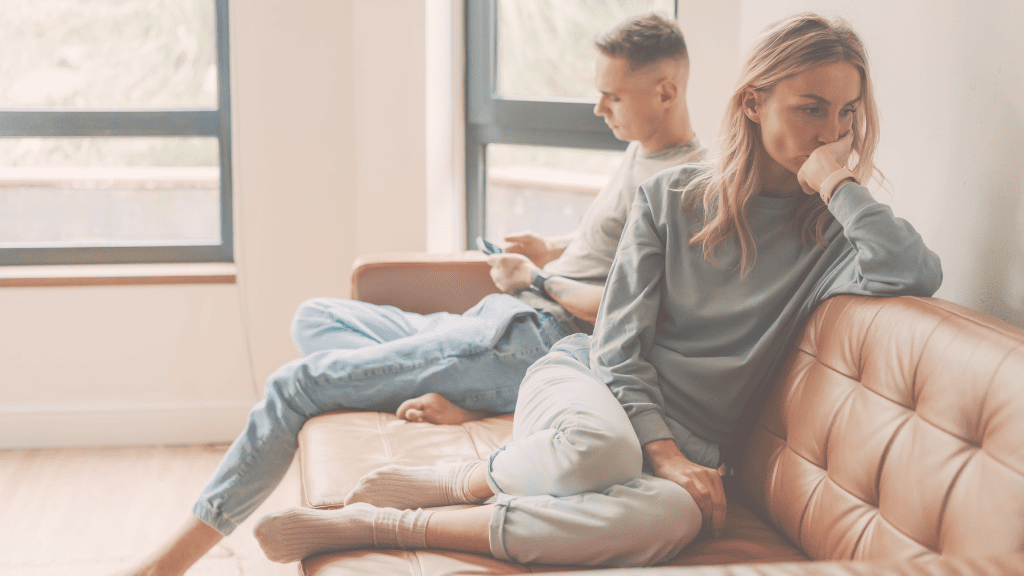 How Your Attachment Style in Relationships Lead to Divorce & 4 Ways to Create a More Secure Attachment Style | MCD couple sitting on a brown sofa, man is playing on his phone, woman is kneeing on the sofa, turned away from him, looking worried and holding her chin with her hand.